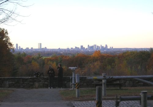 View of a city skyline from a hilltop, surrounded by autumn foliage and people enjoying the scenery.