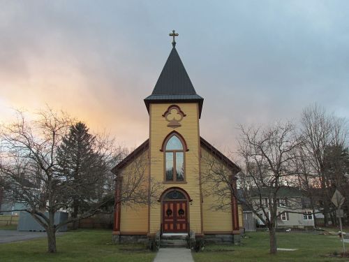 A yellow church with a steeple and cross, surrounded by bare trees, against a cloudy sunset sky.