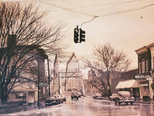 Historic street scene with a traffic light, vintage cars, and a bridge under construction, surrounded by bare trees.