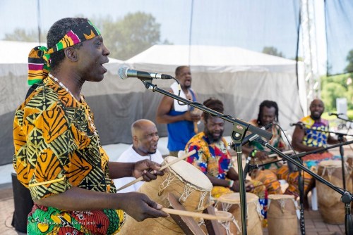 A group of musicians in colorful attire play drums and sing on stage at an outdoor event.