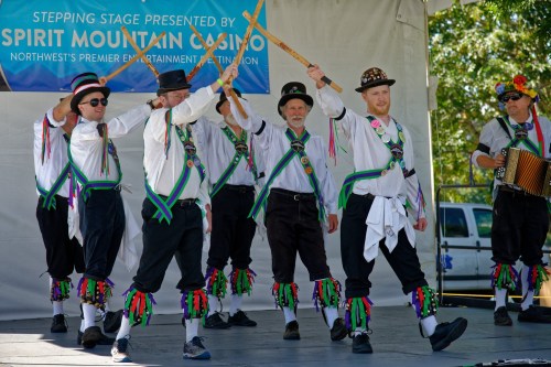 A group of dancers in traditional attire perform with sticks on stage at a festival, with an accordion player nearby.