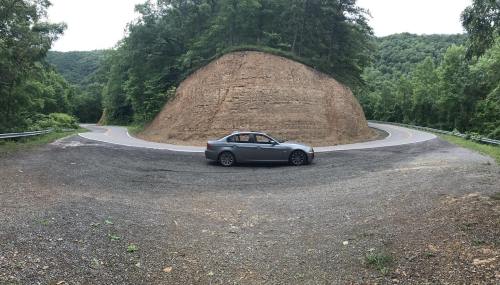 A gray car parked on a winding road beside a large, grassy hill in a forested area.