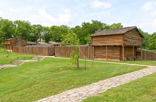 Reconstructed wooden buildings and a stockade surrounded by green grass and trees under a blue sky.