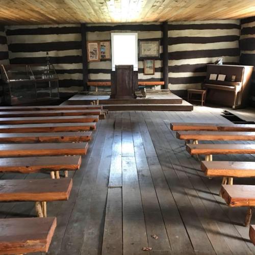 Interior of a rustic wooden room with benches, a podium, and a piano, featuring log walls and a wooden floor.