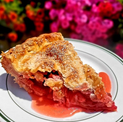 A slice of rhubarb pie on a plate, with a flaky crust and pink filling, set against a backdrop of colorful flowers.