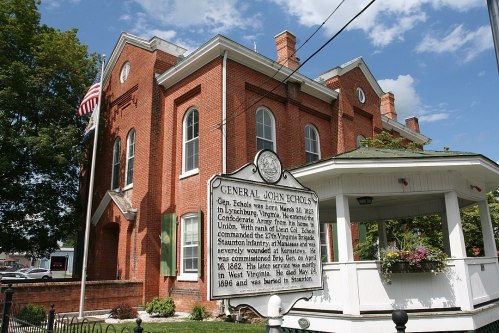 Historic building with a sign for General John Scholl, featuring a gazebo and American flag in the foreground.
