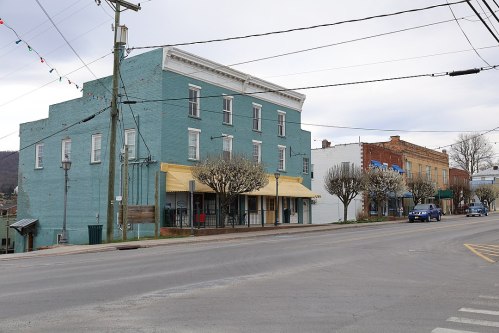 A quiet street scene featuring colorful buildings, flowering trees, and a few cars on a cloudy day.