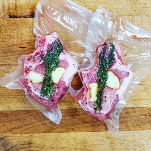 Two vacuum-sealed pieces of meat with herbs and garlic, resting on a wooden surface.