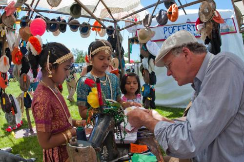 A man assists three girls in traditional attire at a market stall filled with hats and colorful decorations.
