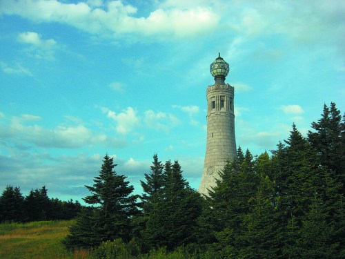 A tall lighthouse stands among green trees under a blue sky with scattered clouds.