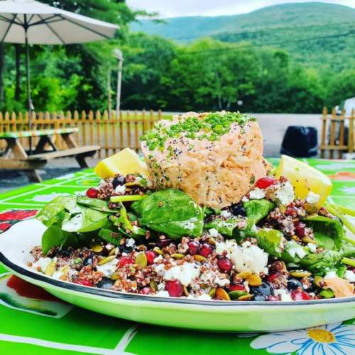 A colorful salad with quinoa, greens, and a large piece of salmon, set outdoors with mountains in the background.