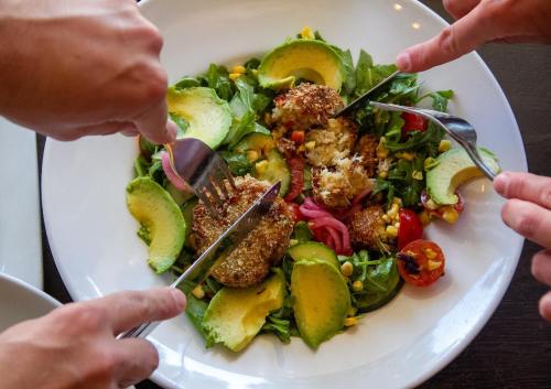 Hands with forks and knives reaching for a vibrant salad topped with avocado, tomatoes, and crispy elements.