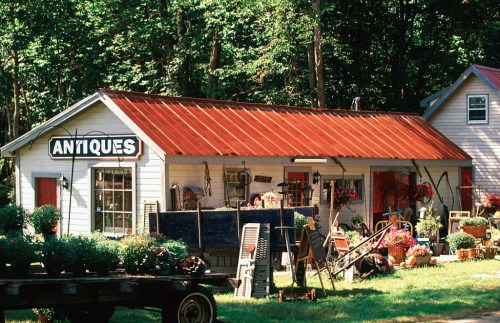 A quaint antiques shop with a red metal roof, surrounded by greenery and filled with vintage items and flowers.