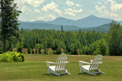 Two white Adirondack chairs on a grassy lawn, overlooking a scenic view of mountains and trees under a blue sky.