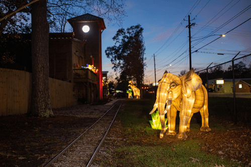 A lit elephant sculpture stands beside a railway track at dusk, with trees and a building in the background.