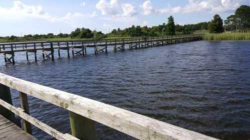 A wooden pier extends over calm water, surrounded by greenery and a blue sky with fluffy clouds.