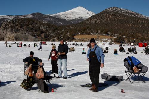People ice fishing on a snowy lake with mountains in the background, enjoying a sunny winter day.