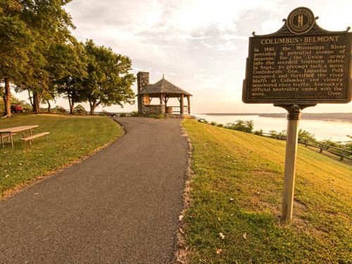 A scenic pathway leads to a gazebo by the river, with a historical marker about Columbus-Belmont in the background.