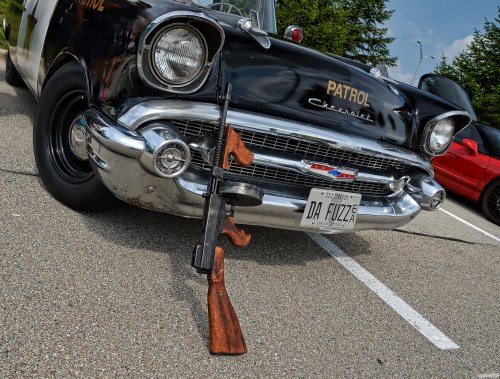 A vintage police car with "PATROL" on the side, featuring rifles leaning against the front and a unique license plate.