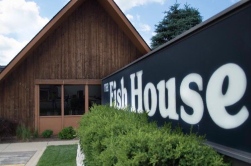 Exterior of The Fish House restaurant, featuring a wooden building and a prominent sign. Green shrubs in the foreground.