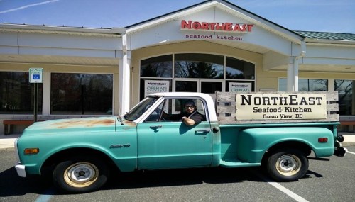 A vintage turquoise pickup truck parked in front of a seafood kitchen with a wooden sign reading "NORTH EAST Seafood Kitchen."