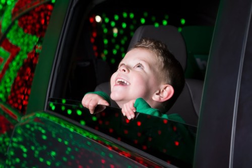 A joyful child leans out of a car window, mesmerized by colorful holiday lights.