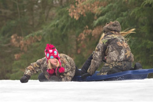 Two children in winter clothing sledding down a snowy hill, one laughing and the other leaning forward on the sled.