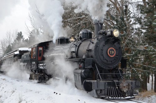 A vintage steam locomotive chugs through a snowy landscape, billowing smoke against a backdrop of trees.