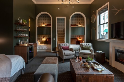 Cozy hotel room with green walls, two beds, a seating area, and modern decor, featuring a fireplace and bar cart.