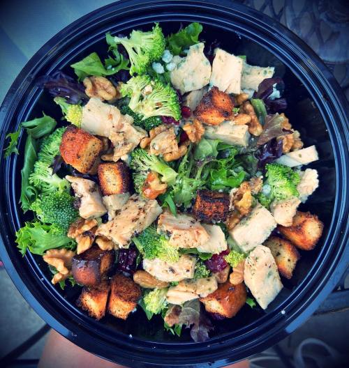 A colorful salad with greens, broccoli, chicken, croutons, walnuts, and cranberries in a black bowl.