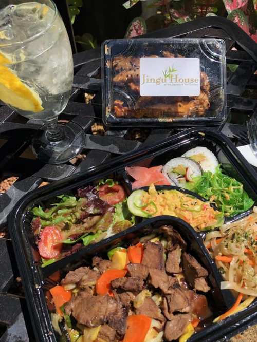 A colorful takeout meal featuring beef stir-fry, sushi, salad, and a drink on a table with greenery in the background.