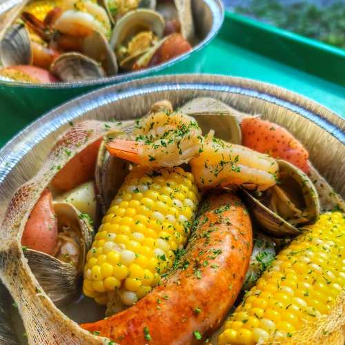 A close-up of a seafood boil with shrimp, corn, sausage, and clams in a metal bowl, garnished with herbs.