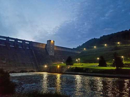 A dam at dusk, illuminated by lights, with a river in the foreground and hills in the background.