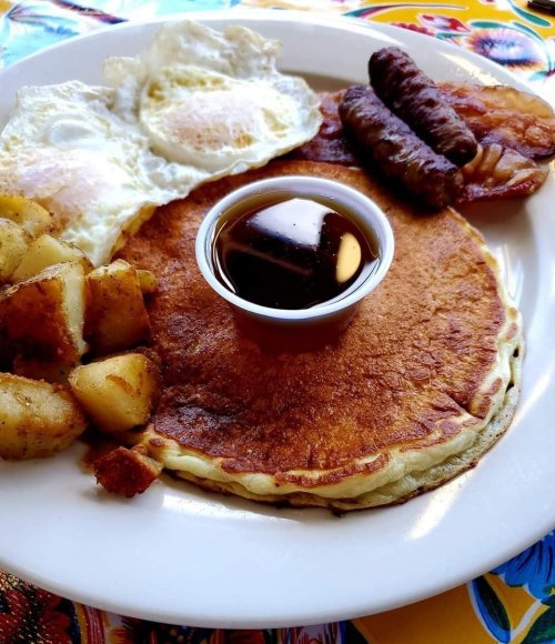 A plate of breakfast featuring pancakes, fried eggs, sausage links, bacon, and seasoned potatoes with syrup.