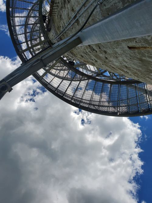 View from below a tall structure, showcasing a circular platform and a blue sky with fluffy clouds.