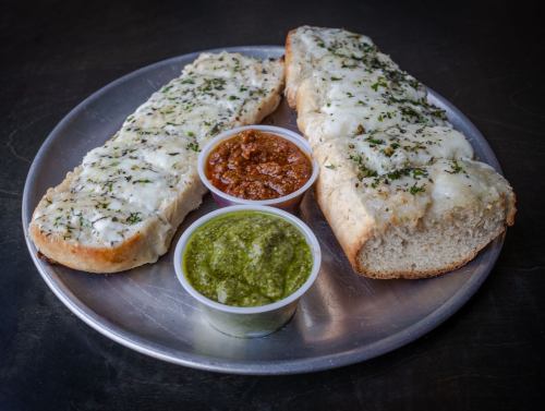 Two pieces of cheesy bread topped with herbs, served with red and green dipping sauces on a metal plate.