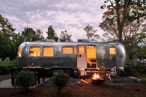 A silver Airstream trailer at dusk, surrounded by trees, with a cozy outdoor seating area and a fire pit.