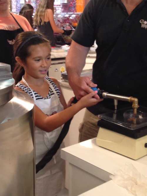 A young girl in an apron assists an adult with a machine in a candy-making shop.