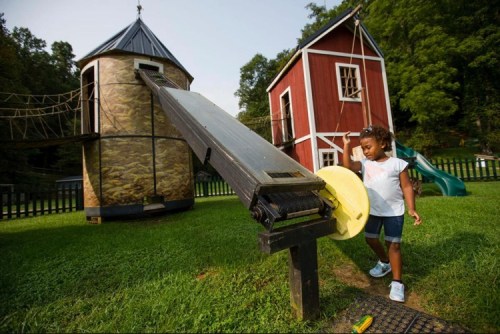 A child plays at a playground featuring a slide, a playhouse, and a grassy area surrounded by trees.