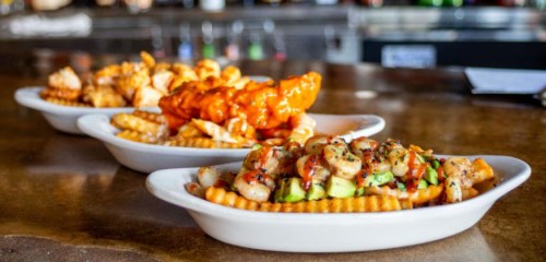 Three plates of crinkle-cut fries topped with shrimp, chicken, and spicy sauce, served on a wooden table.