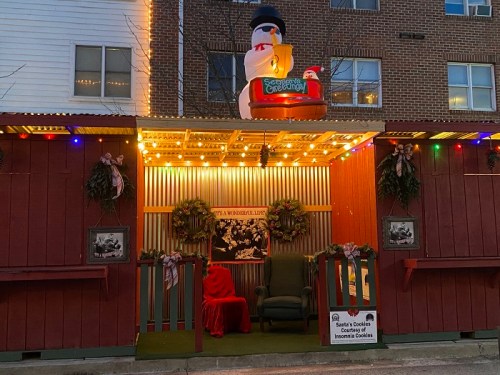 A festive holiday display featuring a snowman, lights, wreaths, and a cozy chair with a red blanket.