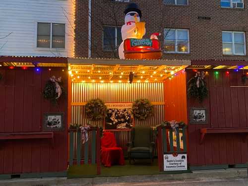 A festive holiday display featuring a snowman, lights, wreaths, and a cozy chair with a red blanket.