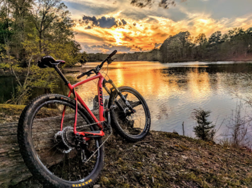 A red mountain bike rests on a log by a calm lake at sunset, with vibrant clouds reflecting on the water.