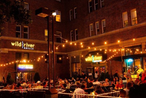Outdoor dining area at night, with string lights and a lively crowd at Wild River Grille. Warm, inviting atmosphere.