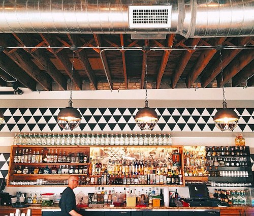 A bar with a wooden ceiling, geometric wall design, and shelves filled with various bottles. A bartender is in motion.