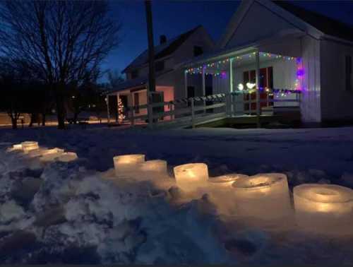 Glowing ice lanterns line a snowy path in front of a house decorated with colorful lights at dusk.