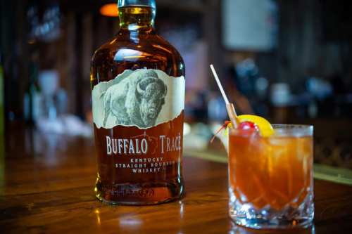 A bottle of Buffalo Trace bourbon next to a cocktail garnished with a cherry and lemon slice on a wooden bar.