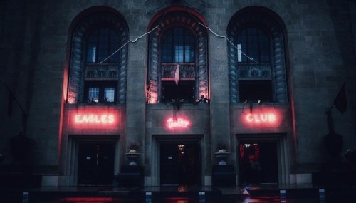 A dimly lit building facade featuring neon signs for "Eagles Club" and "Lounge" with a moody atmosphere.