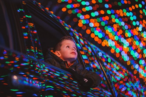 A child gazes out of a car window, surrounded by colorful, blurred holiday lights.