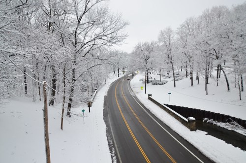 A winding road through a snowy landscape, lined with frosted trees and a small creek beside it.